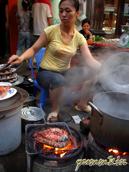 Woman preparing shellfish & grilling over fire in the streets of Hanoi, Vietnam.